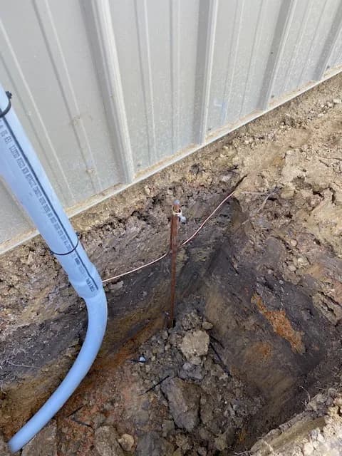 Copper ground rod set in fresh trench at base of shop wall with PVC service conduit running down
